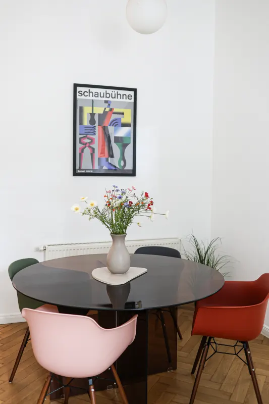 Round dining table with pink, red and green Eames chairs beneath a Bauhaus theatre poster